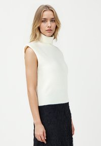 Weißer ärmelloser Rollkragenpullover mit geripptem Saum, der eine figurbetonte Silhouette und eine weiche Textur aufweist, kombiniert mit einem strukturierten schwarzen Spitzenrock.