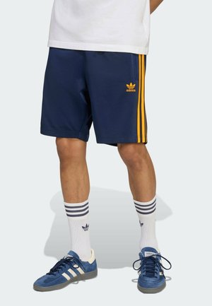 Osoba w granatowych szortach Adidas z żółtymi paskami, białych skarpetkach z niebieskimi paskami, niebieskich butach Adidas i białej koszulce, stoi.