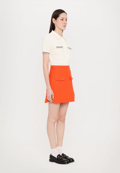 Korte mouwen, romige gebreide polo met gestreepte zakken, gecombineerd met een feloranje minirok met twee flapzakken. Zwarte schoenen maken de look compleet.