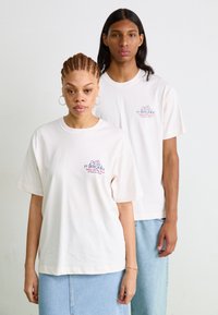 Camisetas oversized de color crema con mangas cortas, que presentan un diseño gráfico en azul y rojo en la zona del pecho izquierdo. Se llevan con faldas de denim.