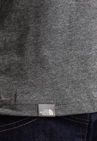 Šedé bavlněné tričko s krátkým rukávem, na kterém se nachází štítek s logem na lemu. Materiál má hladkou texturu s jemným melírováním.