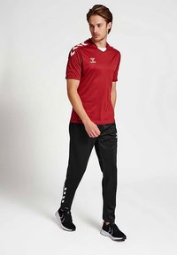 Rood atletisch shirt met korte mouwen en witte accenten op de schouders, gecombineerd met zwarte atletische broek met witte zijstrepen en zwarte sneakers.