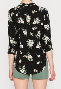 Donna che indossa una blouse nera a maniche lunghe con fiori rosa e pantaloni corti verdi, vista di spalle su uno sfondo semplice.