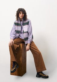 Strukturierte lila Weste mit grünen Streifen über einem lilafarbenen Sweatshirt, kombiniert mit braunen weiten Hosen und schwarzen Wanderstiefeln, sitzt auf Holz.
