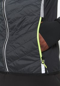 Veste rembourrée noire avec une surface texturée, dotée d'une poche zippée accentuée de jaune fluo et de détails gris réfléchissants.