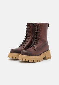 Botas de cuero marrones hasta el tobillo con puntera redondeada, cordones en la parte delantera y cremallera lateral. Suela de goma gruesa y estriada en un color tan contrastante.