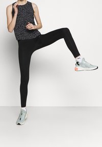 Czarny top bez rękawów w lamparta, w połączeniu z czarnymi legginsami i jasnoniebieskimi butami sportowymi z akcentami, ustawiony z jedną nogą uniesioną.