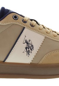 Světlé hnědé tenisky s béžovými a námořnickými akcenty, béžové šňůrky, gumová podrážka a černé logo pikového hráče na bočním panelu.