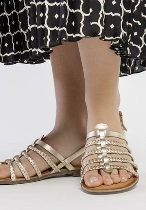 Sandalias doradas con tiras entrelazadas, suela plana, diseño de punta abierta, que cuentan con tiras delgadas y un acabado similar al cuero.
