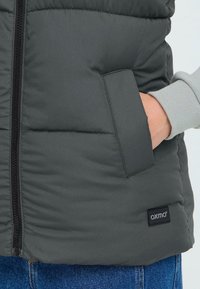 Padded prsluk u tamnosivoj boji s crnim zatvaračem, bočnim džepom i sivim manžetama. Sadrži "oxmo" logotip na donjem rubu.