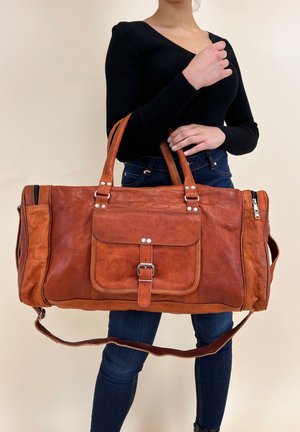Sac de voyage en cuir marron avec une poche frontale, une bandoulière réglable et des accessoires en acier inoxydable. Texture lisse avec des détails de coutures visibles.
