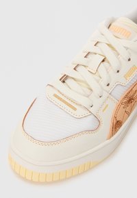 Pantofi sport Puma cu parte superioară din piele crem, țesătură albă nervurată, broderie florală piersică, talpă galbenă și cusături tonale.