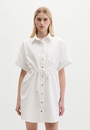 Kobieta w białej sukience koszulowej z krótkim rękawem, z przodu zapinanej na guziki i ze ściągaczem w talii, stojąca na jednolitym jasnym tle.