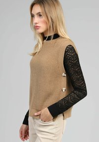 Pull en maille beige avec des manches en dentelle noire, comportant des accents de boutons sur le côté et un col haut. Texture lisse avec un motif côtelé.