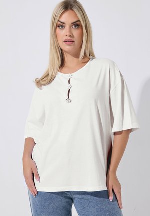 Blonde Frau trägt ein locker sitzendes weißes Oberteil mit drei blumenförmigen Knöpfen und hellblauen Jeans, steht vor einem schlichten Hintergrund.