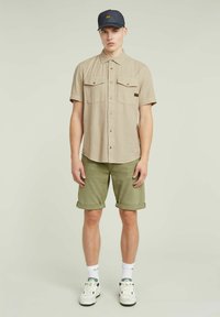 Beige overhemd met korte mouwen en twee borstzakken, gecombineerd met omgeslagen olijfgroene shorts. Een donkere pet en witte sneakers maken de outfit compleet.