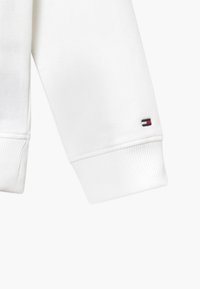 Weißer Sweatshirtärmel mit geripptem Saum, der am Ende ein kleines Logo in Marineblau, Rot und Weiß zeigt. Aus weichem Stoff gefertigt.