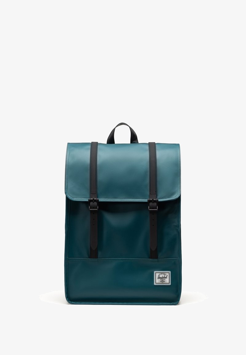 Teal reppu, jossa on kiiltävä pinta, kaksi mustasta nahasta valmistettua olkahihnaa ja läppälynkki, jossa on metalliset soljet. Mukana etutasku.