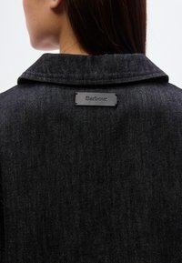 Μαύρο τζιν μπουφάν με διπλωμένο γιακά και ανάγλυφη μαύρη ετικέτα με το όνομα της μάρκας στην πλάτη. Υφασμένο ύφασμα με διακριτική γυαλάδα.