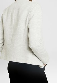 Veste gris clair avec un motif en chevrons texturé. Présente une coupe ajustée avec un bas évasé à volant et des épaules structurées.