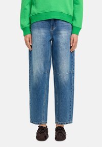 Jasnoniebieskie dżinsy o luźnym kroju, z wyblakłą teksturą i widocznymi detalami szwów. Noszone z zieloną bluzą i ciemnymi butami.