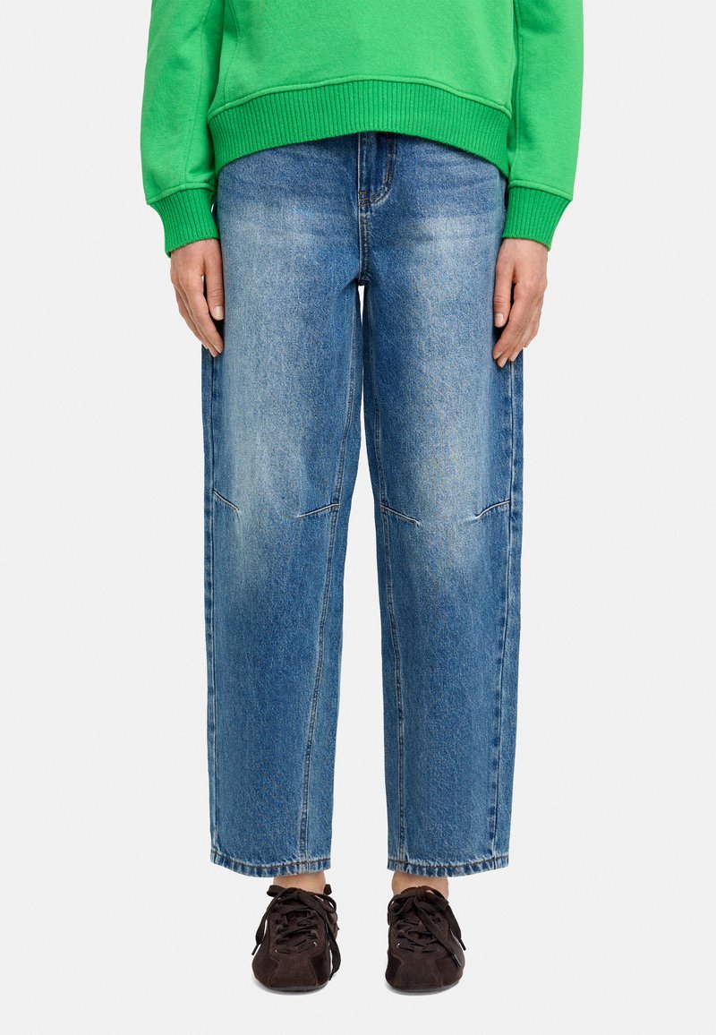 Jasnoniebieskie dżinsy o luźnym kroju, z wyblakłą teksturą i widocznymi detalami szwów. Noszone z zieloną bluzą i ciemnymi butami.