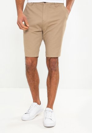 Mand iført beige shorts og hvide sneakers står med den ene hånd i lommen mod en ensfarvet hvid baggrund.