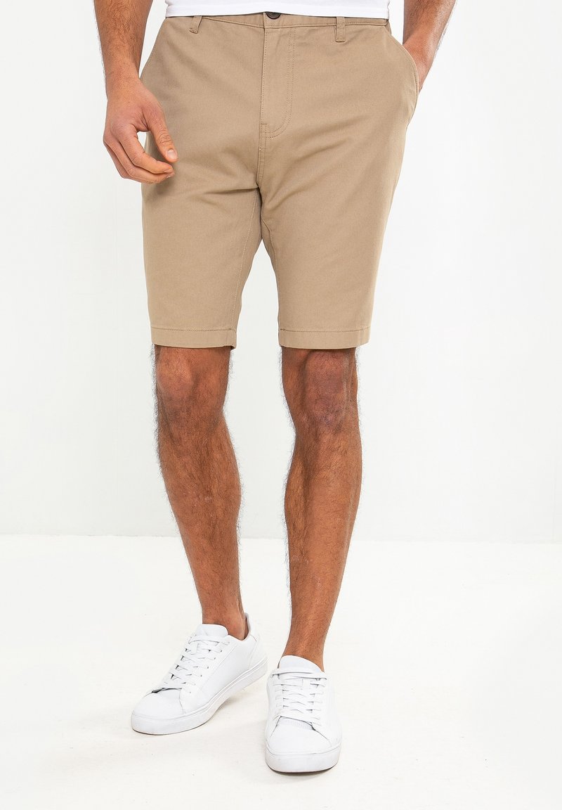 Homme portant un short beige et des baskets blanches, debout avec une main dans la poche sur un fond blanc uni.