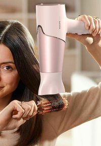 Asciugacapelli Philips, 2000W, corpo rosa con dettagli in argento, tenuto sopra una spazzola rotonda in legno. Capelli scuri, lisci e dritti in primo piano.