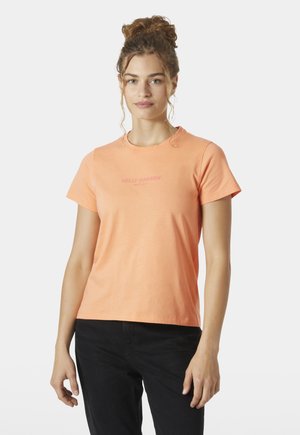 Jonge vrouw met krullend haar in een knot, die een perzikkleurig Helly Hansen T-shirt met korte mouwen en een zwarte broek draagt, staand tegen een witte achtergrond.