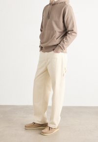 Beige hoodie med framficka, i kombination med krämfärgade byxor och ljusbruna skor. Välpassad med en enkel design och mjuk textur.