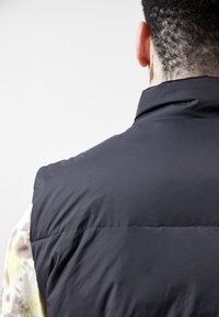 Czarna pikowana kamizelka z wysokim kołnierzem, gładką teksturą i niewidocznymi szwami z tyłu. Noszona na wzorzystej koszuli.