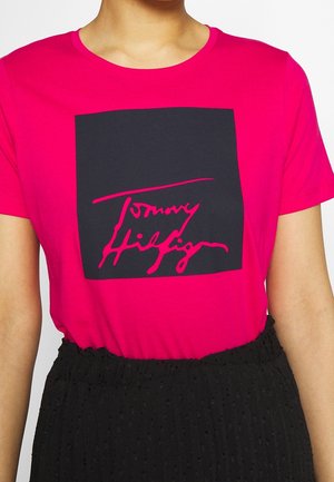 Fuksianpunainen puuvillainen t-paita, jossa on rohkea musta neliökuvio ja valkoista kursiivitekstiä "Tommy Hilfiger". Sileä pinta ja pyöreä kaula-aukko.