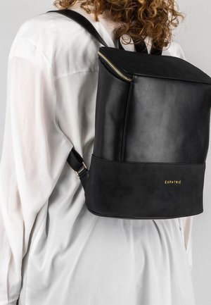 Black leather backpack with a square design, gold zipper, and embossed brand name. Adjustable straps and textured surface details.