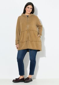Giacca a zip con cappuccio marrone, con una finitura testurizzata, caratterizzata da un dettaglio di vita arricciata e due tasche frontali, abbinata a jeans blu e scarpe nere.