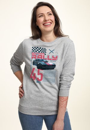 Lächelnde Frau mit langen dunklen Haaren, die einen grauen Sweatshirt mit einem schwarzen Vintage-Rennwagen, einer schwarz-weiß karierten Flagge und dem Text "Gotham Rally 45" trägt.