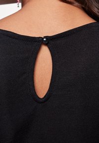 Haut en tricot noir avec une découpe en clé au dos, fermé par un bouton noir rond. Texture lisse avec un design ajusté.