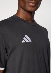 Μαύρο κοντομάνικο T-shirt από μαλακό ύφασμα, με εμφανές λευκό λογότυπο Adidas στο αριστερό στήθος. Απλός σχεδιασμός στο ντεκολτέ.