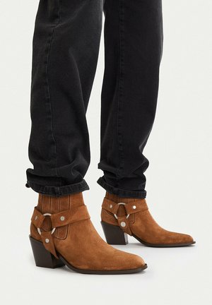Hnědé semišové kotníkové boty se stříbrnými kruhy a pásky, nošené s černými ohrnutými džíny na bílém pozadí.
