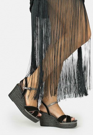 Picioare în sandale cu platformă negre țesute, cu barete la glezne, parțial acoperite de o rochie lungă neagră cu franjuri, pe fundal alb.