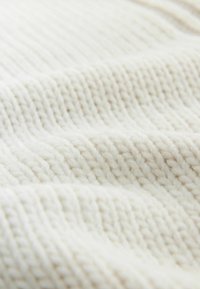 Vastag kötött anyag törtfehér színben, texturált, bordázott mintázattal, puha, vastag fonalakból. A felület finom fényváltozásokat tükröz.