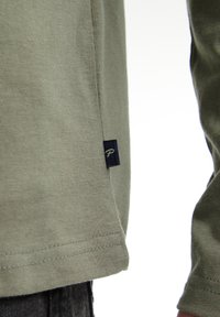 Cămașă verde măsliniu cu mânecă lungă, dintr-un material textil din bumbac, având un etichetă bleumarin cu un emblemă albă pe tiv. Marginile cusute sunt vizibile.