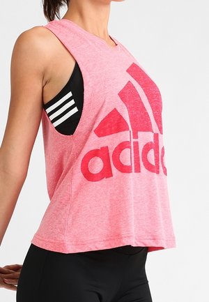 Kobieta nosząca luźny różowy bezrękawnik Adidas na czarnym sportowym biustonoszu z białymi paskami, stojąca z rękami lekko cofniętymi.