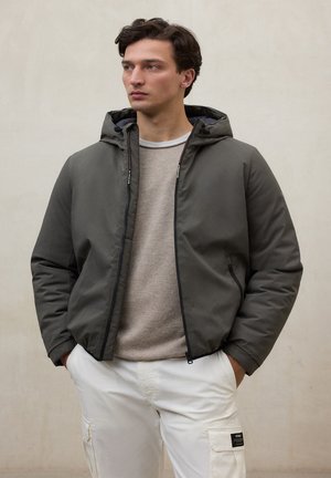 Joven con chaqueta gris con capucha sobre un suéter beige y pantalones cargo blancos, de pie con las manos en los bolsillos, mirando hacia la izquierda.