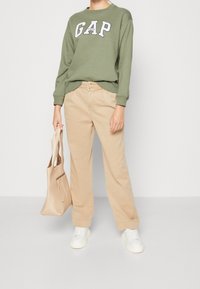 Felpa verde con logo "GAP", pantaloni beige a gamba larga, sneakers bianchi e una borsa a tracolla leggera beige. Tessuto morbido e vestibilità casual.