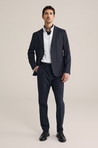 Marineblauw pak met een gestructureerde afwerking, wit overhemd en zwarte schoenen. Voorzien van een sjaal met paisley-patroon rond de hals. Klassiek model.