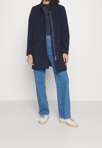 Mörkblå ullkappa med hög krage, dragkedjestängning och två framfickor, bärs över en mörk överdel och ljusblå jeans. Skor med gummisula.