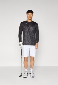 Svart langermet skjorte med abstrakte mønstre, hvite shorts, golfkøl, og grå joggesko. Modellen har på seg en svart caps og golfhansker.