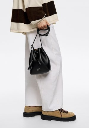 Black leather bucket bag with drawstring closure, logo in white, held in hand. Worn with white wide-leg pants and tan lace-up shoes.