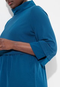 Teal sininen mekko, jossa on korkeakauluksinen malli ja kolmen neljänneksen hihat, valmistettu pehmeästä kankaasta, rento istuvuus ja taskuyksityiskohta.